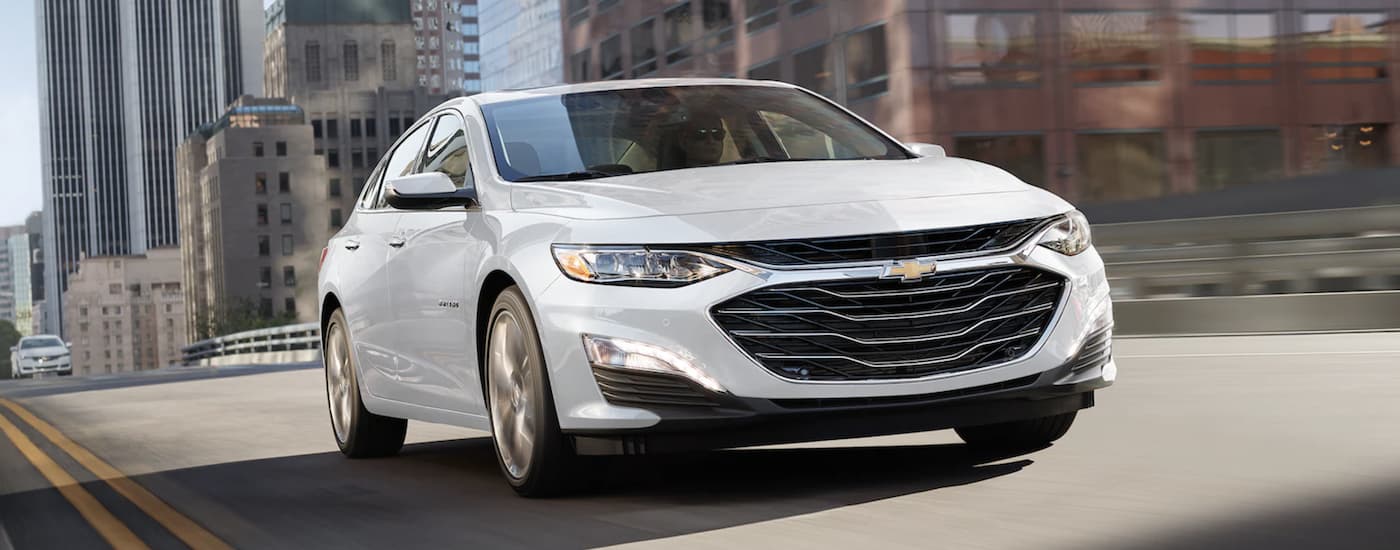 A white 2021 Chevy Malibu is shown from the front driving through a city.
