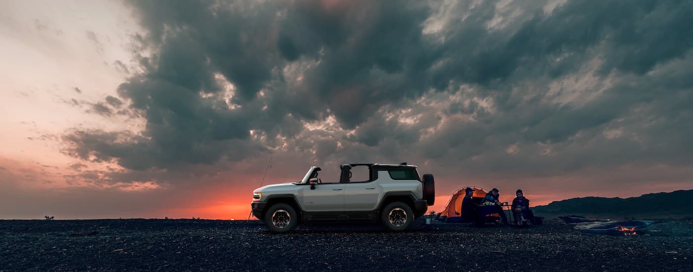 A white 2024 GMC Hummer EV is shown from the side parked near a campsite at sunset.