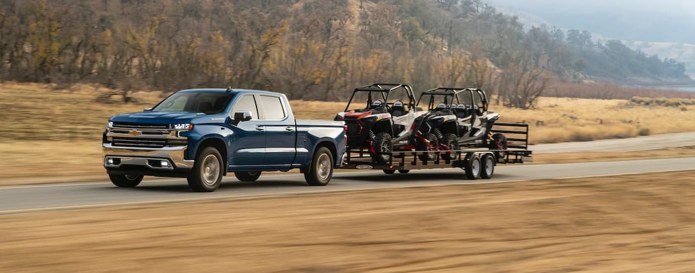 A blue 2020 Chevy Silverado 1500 is shown towing a trailer of UTVs on a highway.
