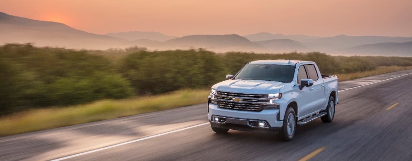 One of the most popular vehicles in pre-owned luxury truck sales, a white 2021 Chevy Silverado 1500 High Country, is shown driving on a tree-lined road.