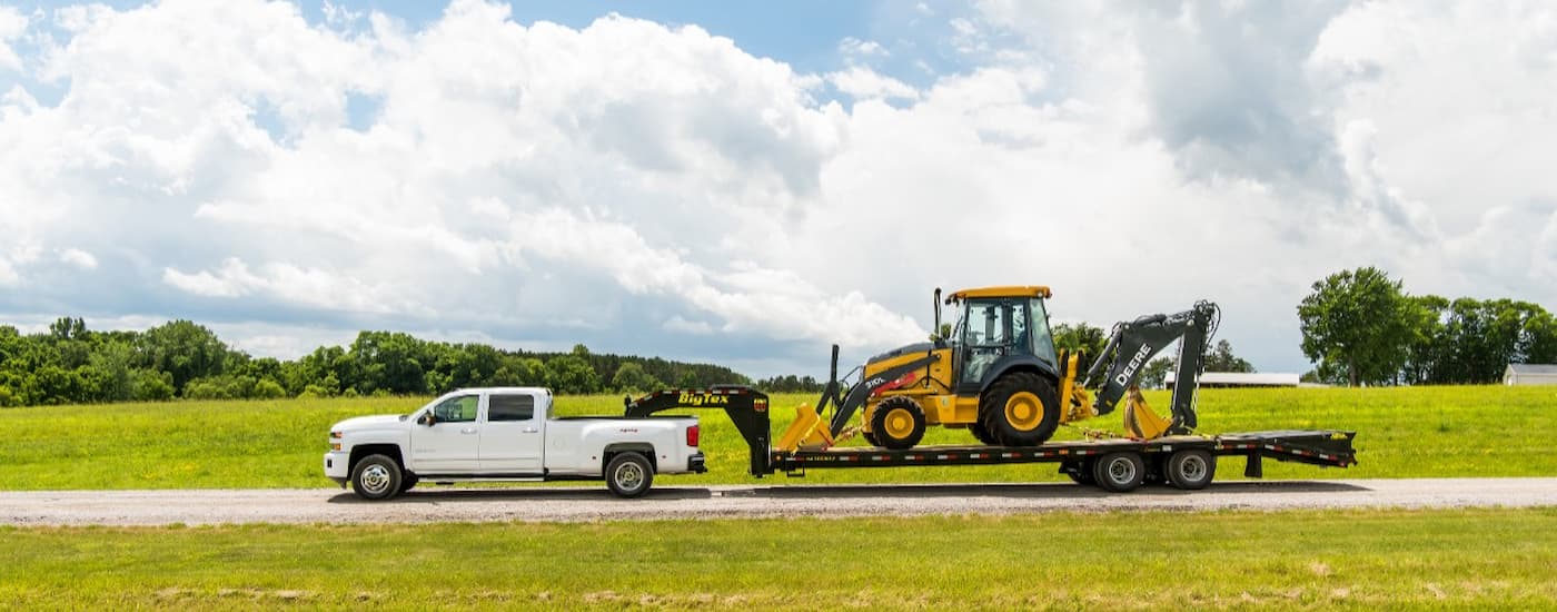 A white 2019 pre-owned Chevy Silverado 3500HD is shown from the side towing heavy machinery. 